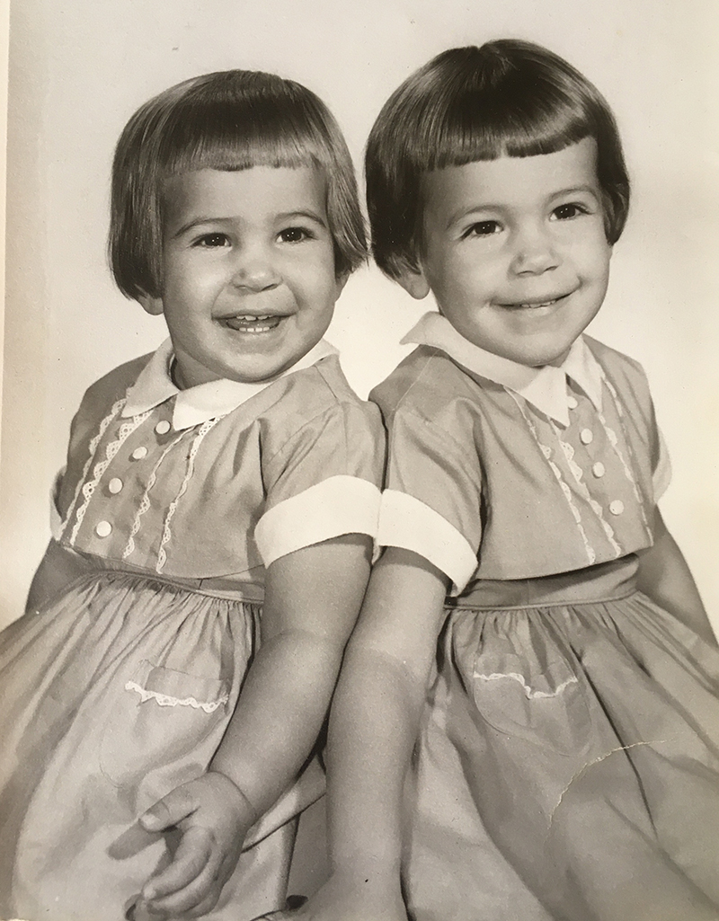Two girls are smiling in a sepia-toned photo. They are wearing identical dresses and have identical cropped haircuts.