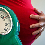 Pregnant woman holding a bathroom-style scale in one hand while cupping her other hand over her stomach.
