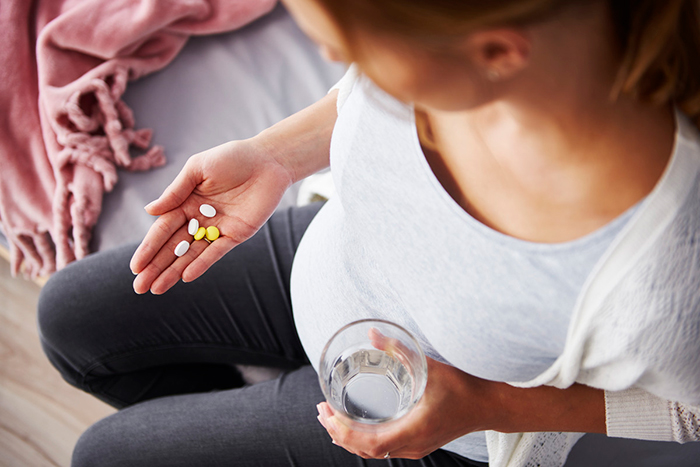 A pregnant person holding medicine tablets in one hand and a glass of water in the other. 