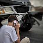 Teenage boy, sitting on curb, talking on a cell phone, in front of a car with front end collision damage.