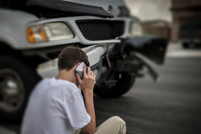 Teenage boy, sitting on curb, talking on a cell phone, in front of a car with front end collision damage.