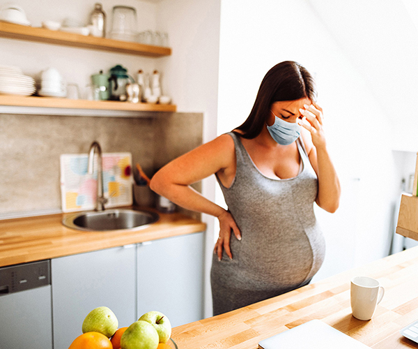 A pregnant woman wears a facemask and appears visibly tired in her kitchen.