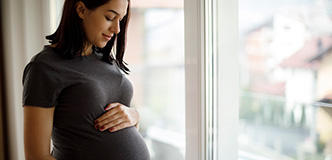 Pregnant person with their hands on their belly, standing in front of a window.