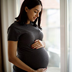 Pregnant person with their hands on their belly, standing in front of a window.