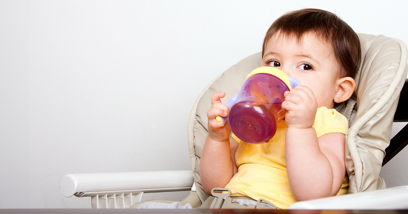 Small child in high chair drinking juice from a sippy cup.