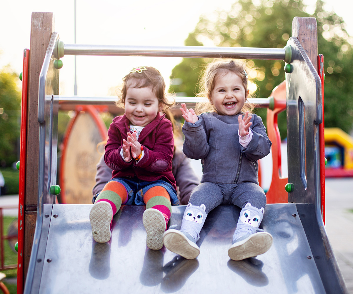 Two toddler girls smile and clap side-by-side at the top of a wide metal slide.