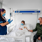 Child lying in hospital bed, concerned parent at bedside. Health care worker writing on clipboard. All are masked.