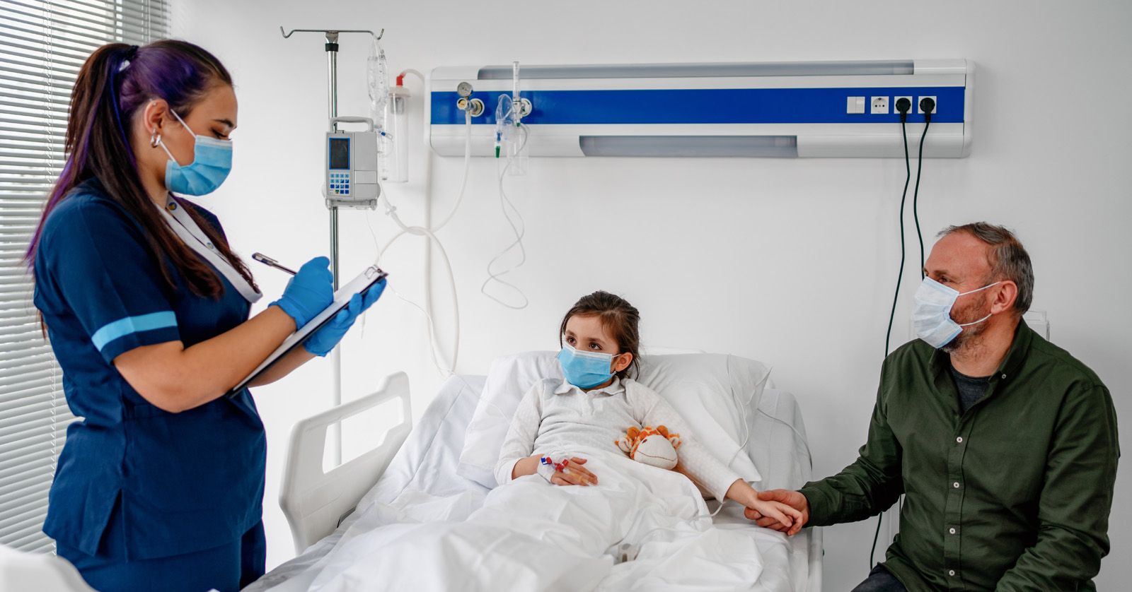 Child lying in hospital bed, concerned parent at bedside. Health care worker writing on clipboard. All are masked.