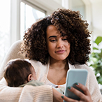 A mother holding an infant and looking at a smartphone.