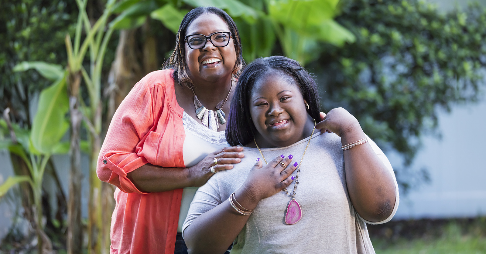 A young woman with Down syndrome smiles with her grandmother. Both are standing outside in front of a tree.