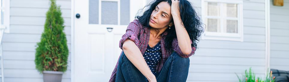 A woman sits on the steps of a deck outside a house with her knees drawn up close to her body. She rests her head in the palm of one hand as she stares into the distance. 