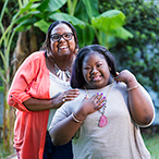 A young woman with Down syndrome smiles with her grandmother. Both are standing outside in front of a tree.