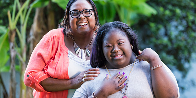 A young woman with Down syndrome smiles with her grandmother in an outdoor setting.