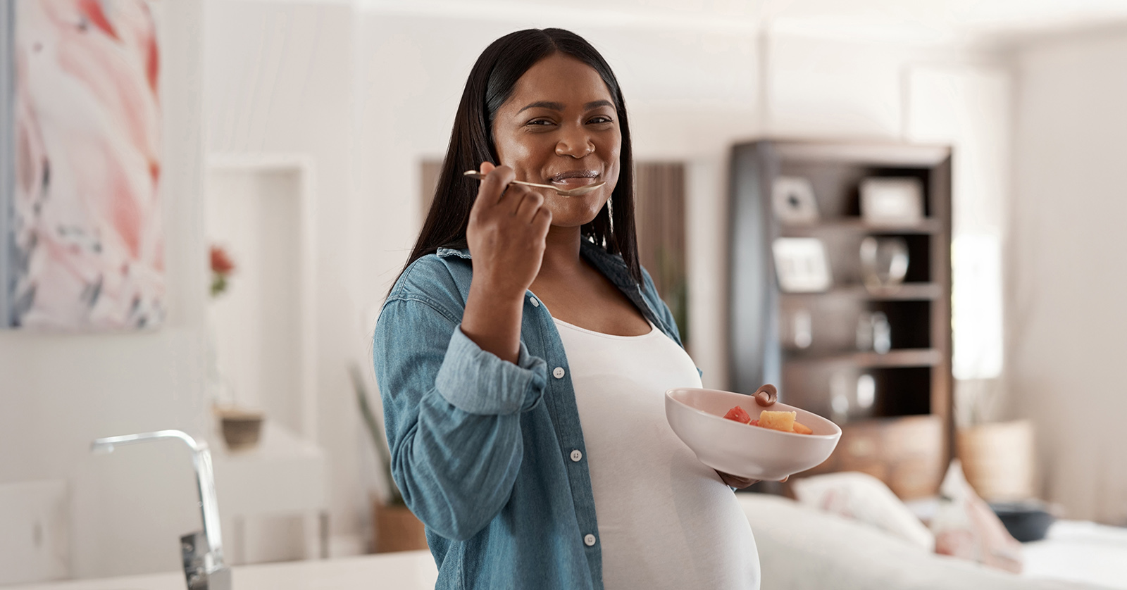 Smiling pregnant woman eating a bowl of multicolored fruit.