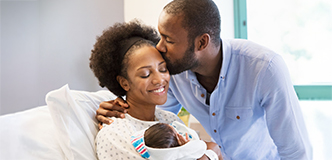 A man kisses a woman on the forehead as she smiles down at the baby she holds in her arms.