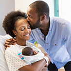 A man kisses a woman on the forehead as she smiles down at the baby she holds in her arms.