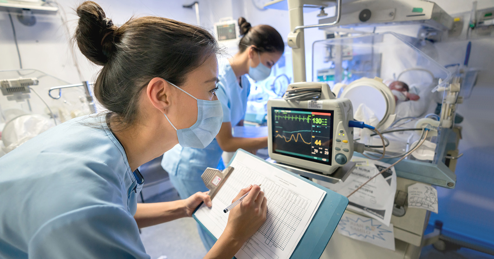 Technicians taking notes from screen monitor of a preterm infant in an isolette.