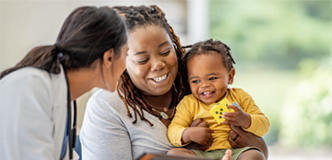 A mom and baby are smiling while a health care provider with a white coat, stethoscope, and electronic tablet looks at them.