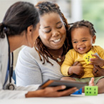 A mom and baby are smiling while a health care provider with a white coat, stethoscope, and electronic tablet looks at them.