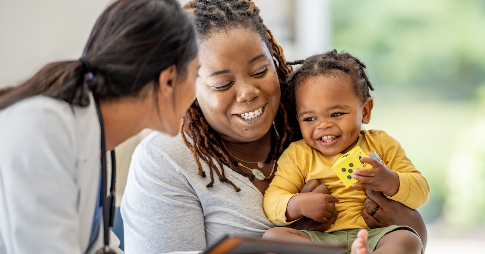 A mom and baby are smiling while a health care provider with a white coat, stethoscope, and electronic tablet looks at them.