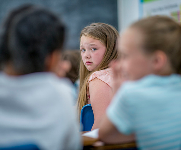 A child sitting in a classroom glances over her shoulder at two classmates who are whispering to each other.