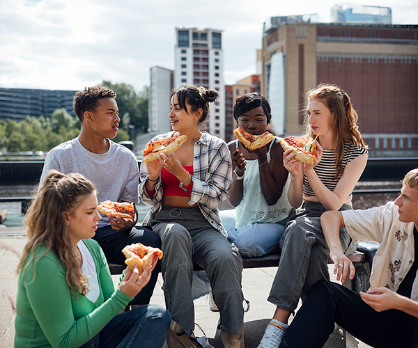 A diverse group of teenagers are sharing a pizza outdoors in a city environment.