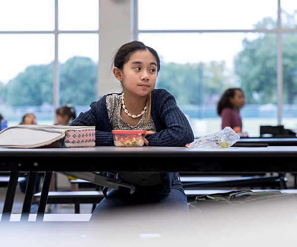 A young teenager sits alone in the cafeteria with a sad look on her face.