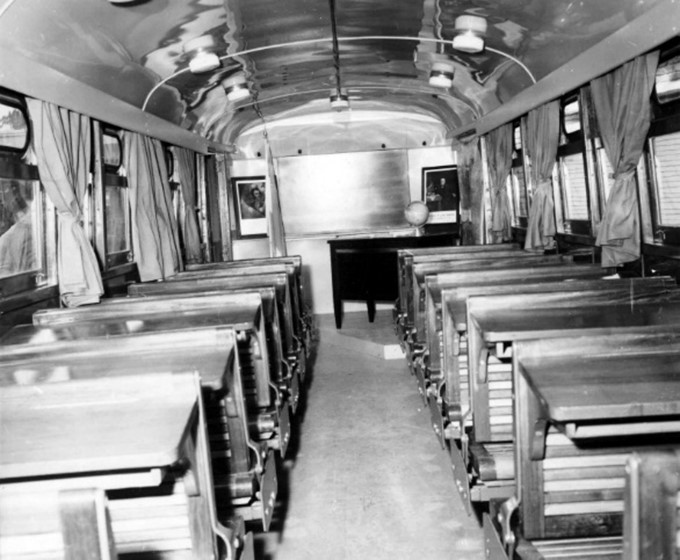 The view from the back of the converted trolley classroom. Rows of benches and tables line both sides, tied curtains frame each window, and a teacher’s desk sets at the front of the trolley.