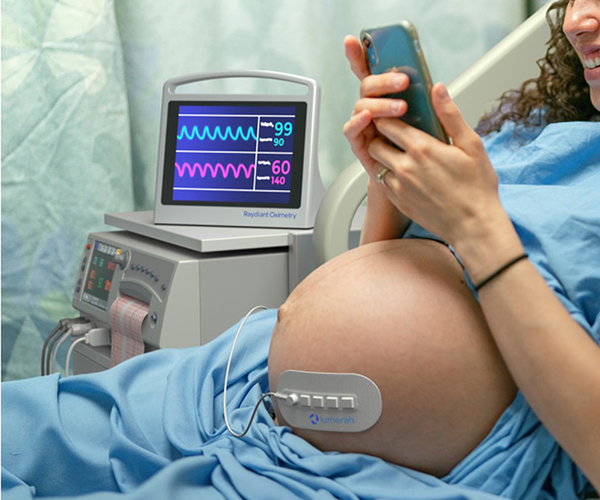 A pregnant women in a healthcare setting has the Lumerah oximeter on her belly while she uses a smartphone.