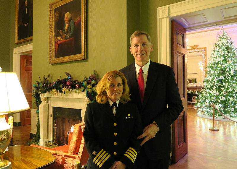 A couple smiles for the camera inside a green room with a fireplace. Dr. Rouault is wearing her Public Health Service uniform. There’s a Christmas tree and decor on the mantle.