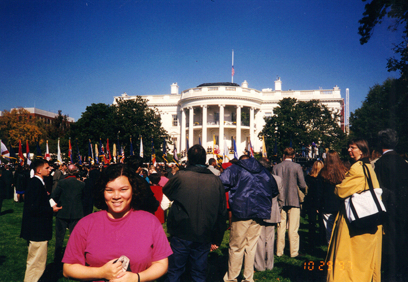 Young woman with dark hair and a pink shirt poses for a photo in front of the White House. A sizable crowd of people can be seen behind her.