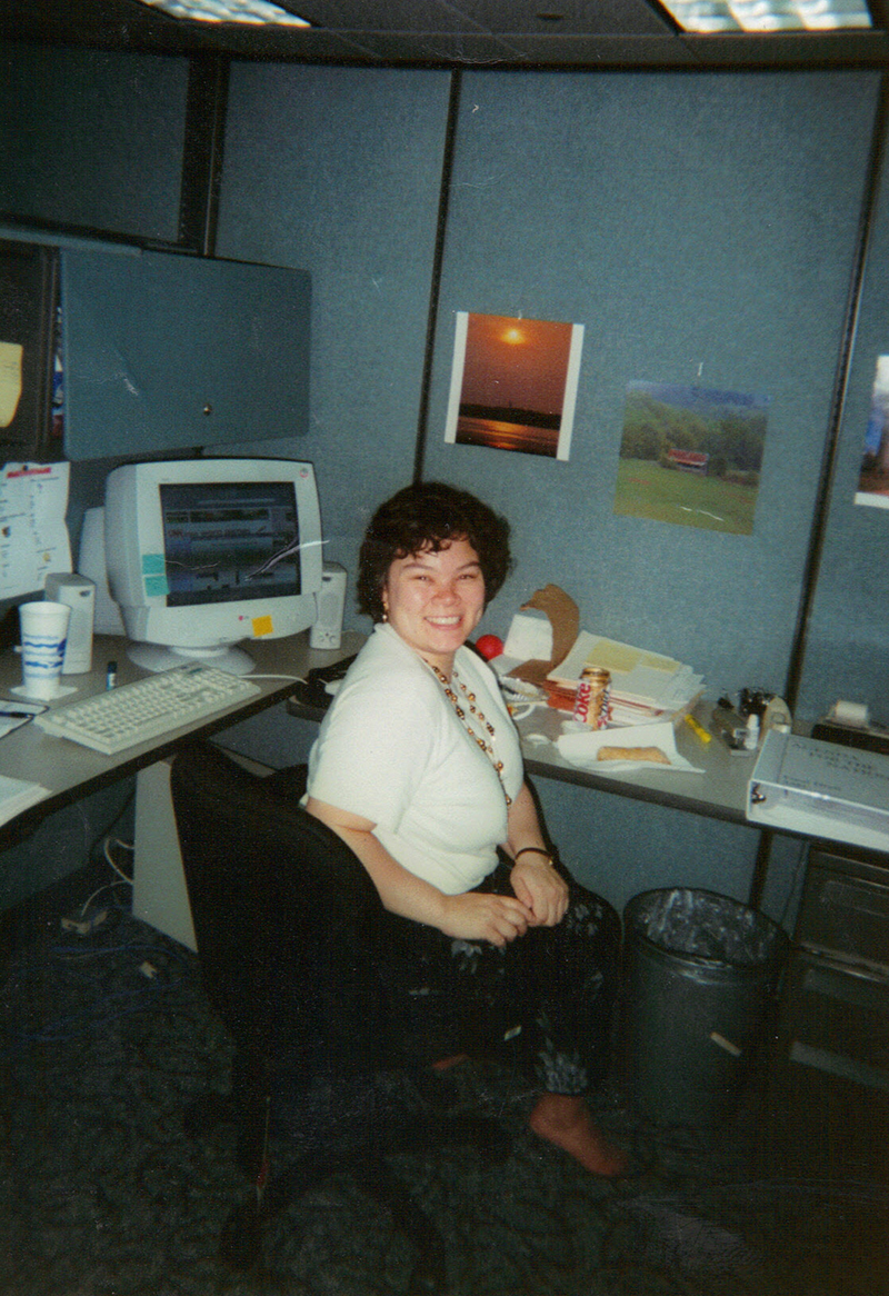 Woman sitting in an office cubicle. An older CRT computer monitor is on the desk behind her and two scenic pictures are pinned to the wall above her workspace. 