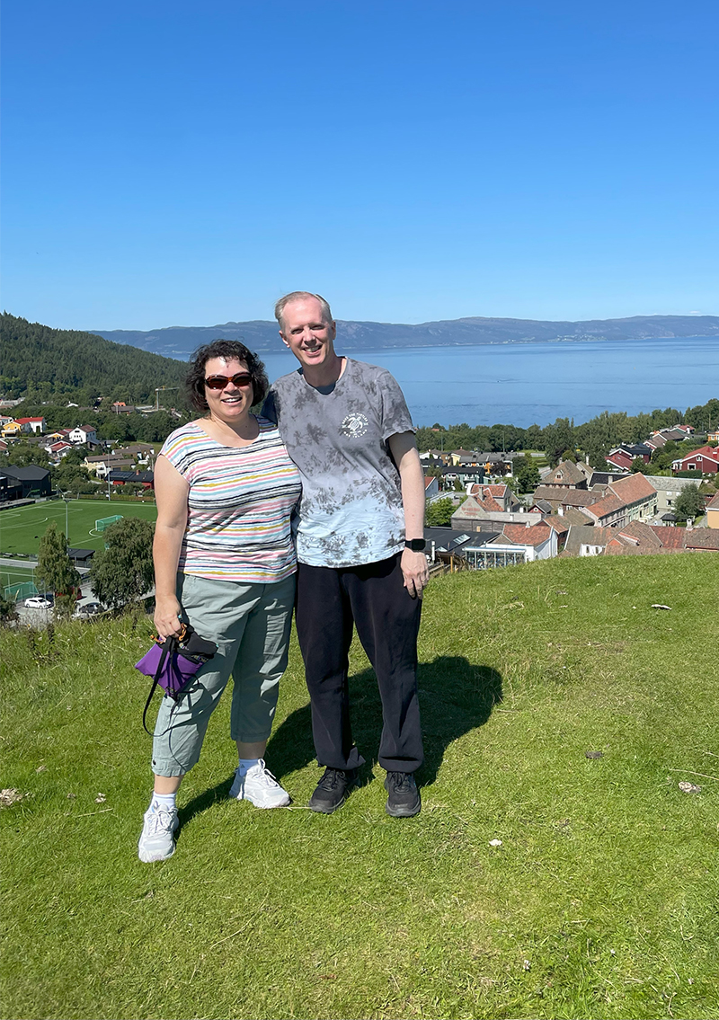 A woman and a man stand together on a green hillside. They are dressed in summer attire. She wears sunglasses. A small village rests below them and water and a mountain line are in the background. 