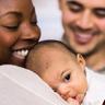 A smiling woman holds a baby as a smiling man looks on in the background.